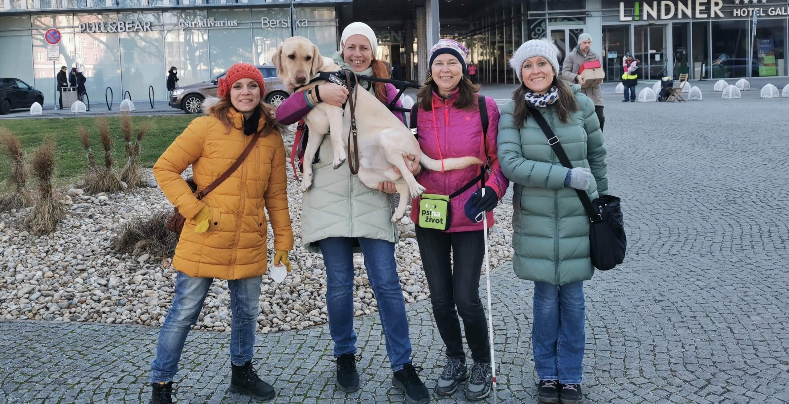 Pred obchodným domom stoja na chodníku 4 vysmiate ženy - členky skúšobnej komisie, pričom jedna drží na rukách žltého labradora v postroji vodiaceho psa.